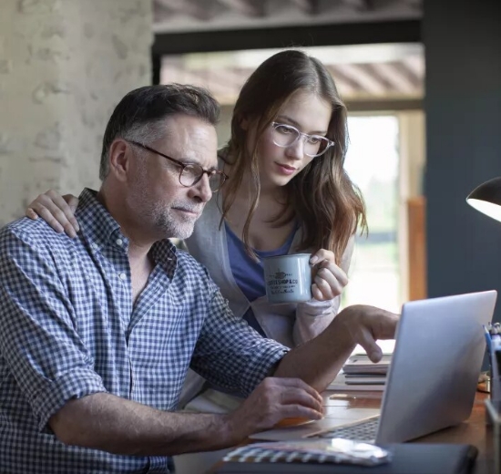 Mann mit Arbeitsplatzbrille sitzt vor Bildschirm. Über seine Schulter gebeugt schaut eine Frau mit Brille und Kaffeetasse ebenfalls mit in das Laptop.